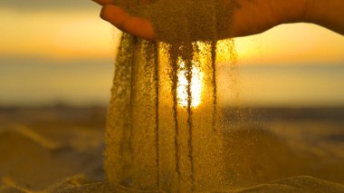 Yakın: Kuru kum küçük parçacıklar nazik kimliği belirsiz bir kadın'ın parmakları arasında ve güzel kıyısında geri düşer. Güzel yaz günbatımı kum plaj ve elleri kuru Kum eleme kadın aydınlatır.