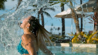 Yakın: Beyaz kız şakacı saçlarını geriye doğru güneşli egzotik havuzda çevirir. Neşeli genç kadın onu saç geri ve spreyler su damlacıkları tropikal havuzu genelinde sıcak yaz gününde parlak kamçı.