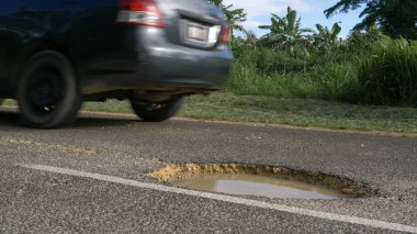 Düşük açılı, Close Up: Gri araba üstünde egzotik adada uzaktan beton yol ağzı açık bir su dolu çukur evades. Yağmur ormanları ile önde gelen bozuk asfalt sokağa tehlikeli bir çukur geçmiş araba kullanıyor.