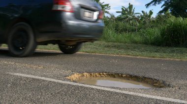 LOW ANGLE, CLOSE UP: Gray car evades a gaping water filled pothole on remote concrete road on exotic island. Car drives past a dangerous hole in the damaged asphalt street leading through rainforest. clipart