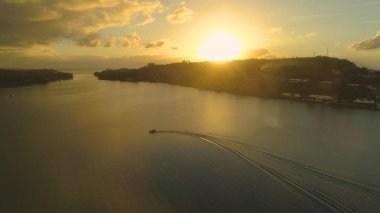 AERIAL, LENS FLARE: Cool unknown wakeboarder does sharp turns on calm river while being tugged by speeding boat on specatcular summer evening. Extreme sportsman carving on wakeboard at golden sunrise.