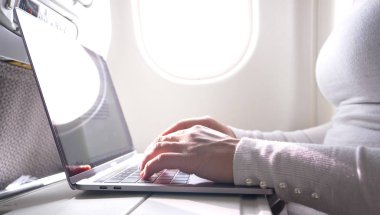 CLOSE UP: Unknown young Caucasian businesswoman types on her high-tech laptop. Bright sunrays shine through white airplane window and on gentle female fingers writing on smooth computer keyboard.