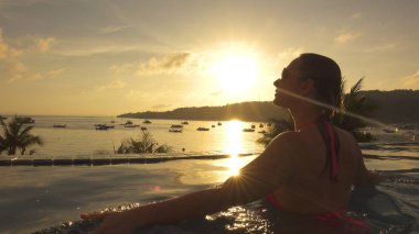 Kadar yakın, Lens Flare: Genç beyaz bir kadın sonsuzluk havuzu kenarına güzel tropikal günbatımında yürür. Genç kadın havuzundan Koyu'ndaki görünümlü Parlatıcı pitoresk altın sabah güneş ışınları.