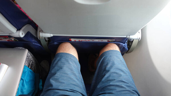 POV: Sitting in an uncomfortable economy class window seat with no leg room. Tourist on long transatlantic flight gets his legs jammed into the seat in front of him. Traveler enduring annoying flight.