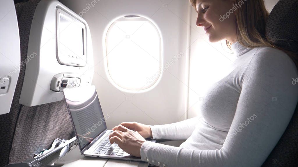 CLOSE UP: Happy Caucasian businesswoman is typing on her high tech laptop during a long flight. Female passenger working on her computer while she flies overseas to an important business meeting.