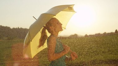 Kadar yakın, Lens Flare: Muhteşem olan kadın sarışın at kuyruğu dansları serin yaz yağmur altında. Beyaz kız sarı bir şemsiye tutan şakacı sakin altın aydınlatılmış kırsal ortamda yağmurda twirls..