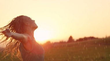 Kopya alanı: Close Up, Lens Flare, gülümseyen genç kadın yağmur altında uzanmış kolları ile döner. Sarışın saçlı beyaz kız şakacı günbatımı çarpıcı dans ediyor. Kadın dansçı yağmurda doğaçlama