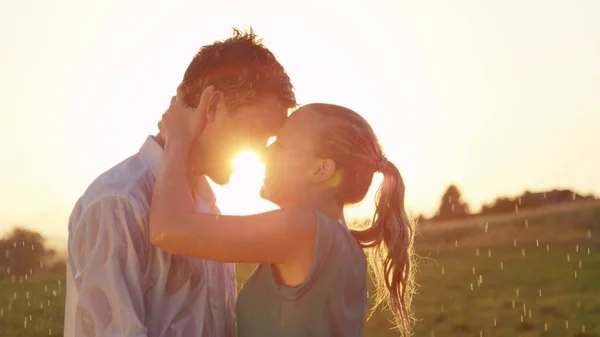 CLOSE UP, LENS FLARE: Beautiful young Caucasian couple kisses and dances in the cool summer evening rain. Carefree boyfriend and girlfriend get wet during their romantic date in the sunny countryside.