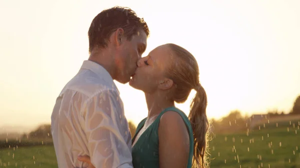 CLOSE UP: Two young lovers kiss in the rain while they dance in the countryside at picturesque summer sunset. Boyfriend and girlfriend kiss during their romantic date on a breathtaking spring morning.