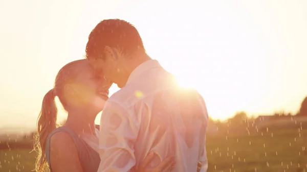 CLOSE UP, LENS FLARE: Embraced young couple dances in beautiful golden lit nature during a spring shower. Caucasian woman and man enjoy a romantic date on a rainy summer morning in the countryside.