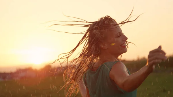 CLOSE UP, LENS FLARE: Attractive woman in pretty green sundress spins in nature on a rainy spring morning. Easygoing blonde girl gets soaking wet while she dances outdoors in the pleasant summer rain.