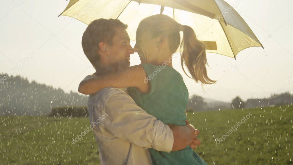 CLOSE UP, LENS FLARE: Cheerful young couple shares a romantic moment while dancing in the summer rain. Lovely Caucasian woman and man spin and look into each other's eyes during fun romantic date.