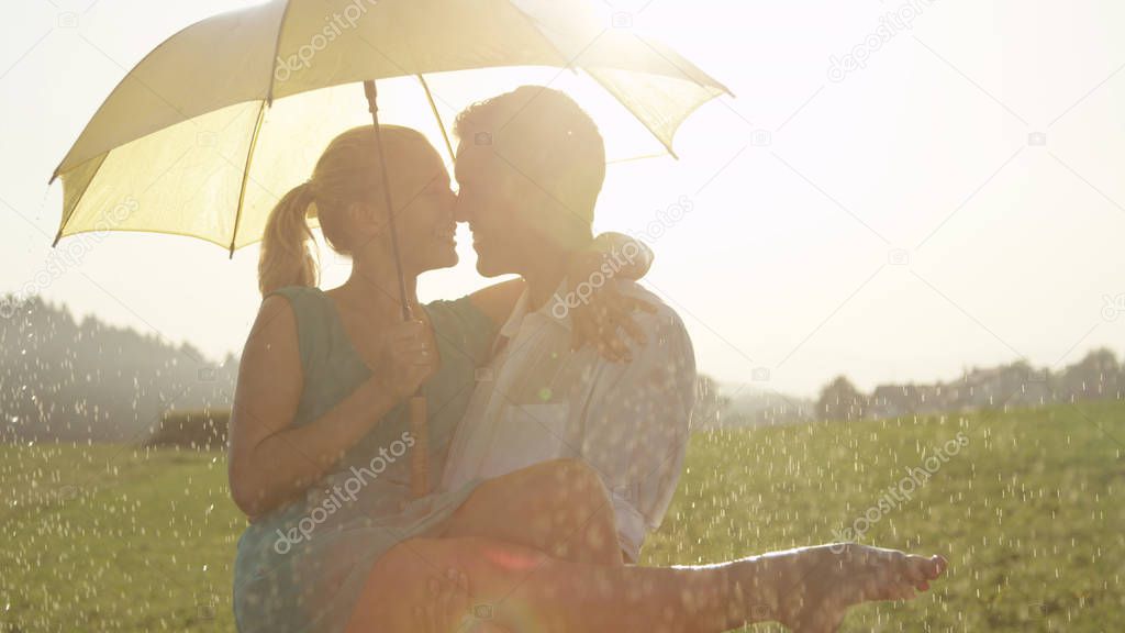 CLOSE UP, LENS FLARE: Embraced young couple is about to kiss while dancing in the cool summer rain. Cheerful man and woman having fun on their outdoor date in the picturesque spring countryside.