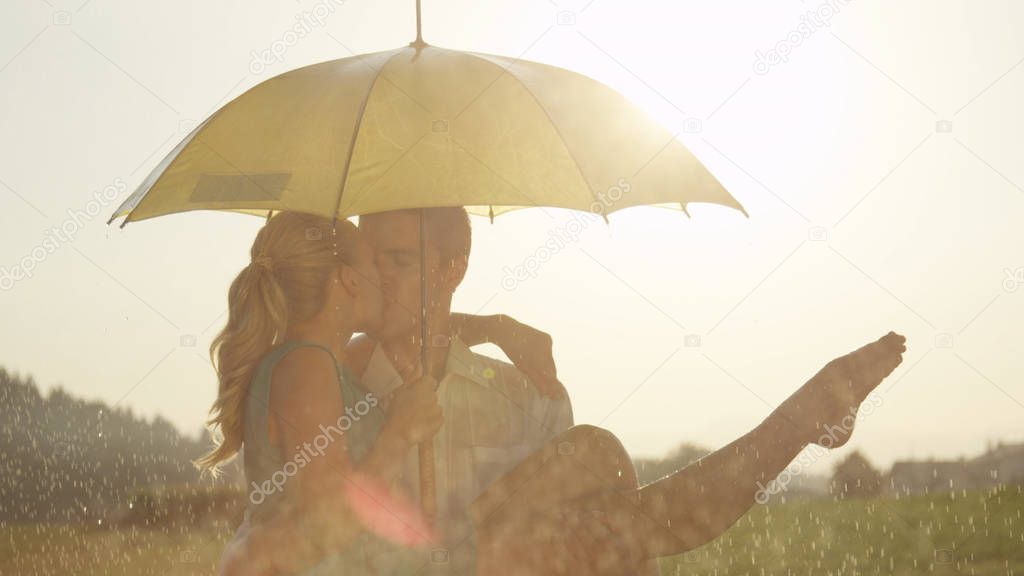 CLOSE UP, LENS FLARE: Joyful man kisses his girlfriend while holding her up and dancing with her in the spring rain. Cute embraced couple kisses under a yellow umbrella during a fun date in nature.