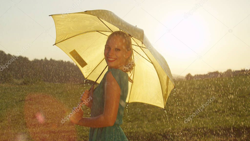 PORTRAIT, LENS FLARE: Smiling blonde woman looks playfully over her shoulder and spins with her yellow umbrella on a refreshing rainy day in the summer. Happy girl dancing alone in the spring rain.