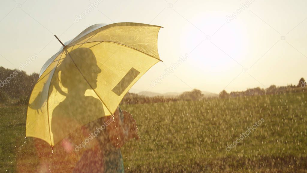 LENS FLARE, CLOSE UP: Unrecognizable young woman hidden behind umbrella cheerfully looks into the distance as refreshing summer rain soaks the green countryside. Carefree girl daydreams on a rainy day