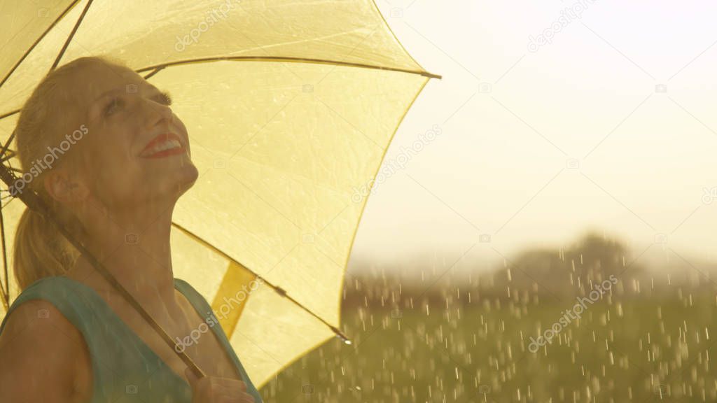 CLOSE UP: Beautiful smiling woman having fun in the refreshing spring rain. Blonde haired girl spins in the rain in a quiet golden lit meadow. Carefree woman in green looking around spectacular nature