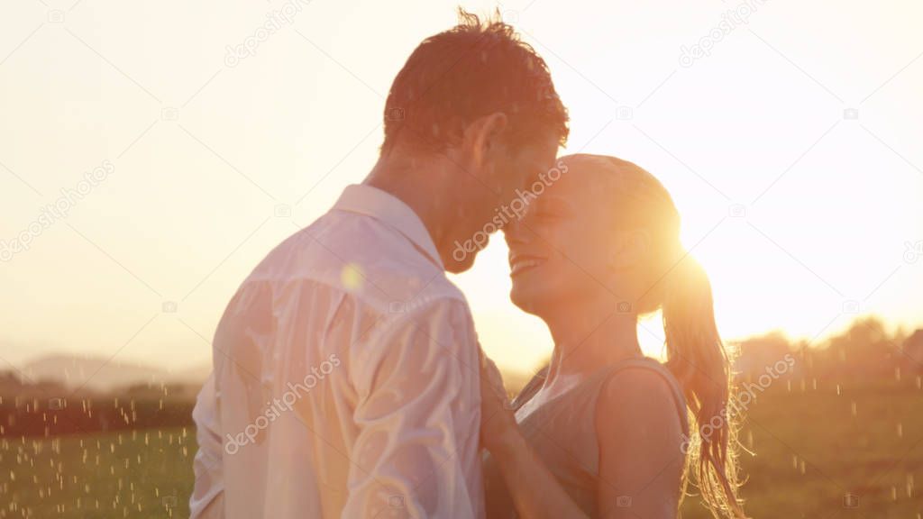 LENS FLARE, CLOSE UP: Cheerful Caucasian couple dancing outside in the rain is about to kiss at breathtaking sunset. Infatuated blonde haired woman gazes into handsome man's eyes before kissing him.
