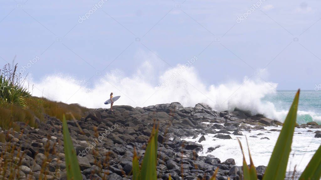 COPY SPACE: Large ocean waves hit the beautiful black rocky beach as surfer girl looks into the distance. Young woman holds a surfboard while she stands still as powerful waves splash across shore.