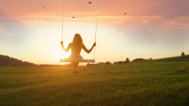 Altın ışık siluet: Beyaz elbiseli huzurlu akşam ağaç salıncakta sallanan tanınmayan kız. Tahta salıncakta oturan ve altın batımını Bayan. Güneş doğarken sallanan genç kadın