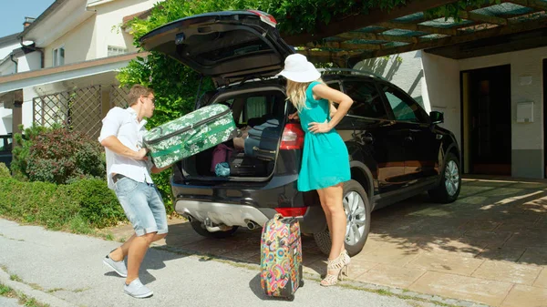 男を持ち上げて重いスーツケース大きな黒い車に妻が彼を監視しながら闘争 快適な郊外の近所の Suv が素晴らしい夏の遠征の準備からの陽気な旅行者 — ストック写真