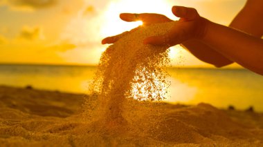 CLOSE UP: Young woman on summer holiday letting coarse sand slip through her fingers. Unrecognizable girl lifts up a handful of white sand over the sunset shining on the exotic beach in Cook Islands.