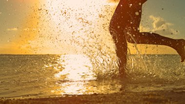 CLOSE UP, LOW ANGLE, SILHOUETTE: Playful girl on holiday runs in the refreshing ocean at sunrise. Unrecognizable young woman goes for a run on the picturesque exotic beach on a sunny summer evening.