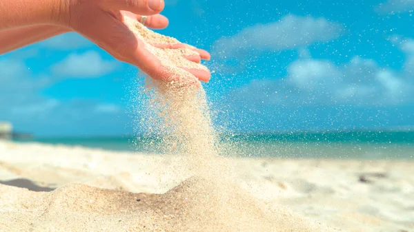 CLOSE UP: Small grains of white sand sift through unrecognizable young woman's fingers. Gentle summer breeze blows away coarse sand in playful female's hand. Girl holds up a handful of white sand.