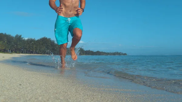 ローアングル 認識できないフィットは男性の観光客はさわやかなエキゾチックな海で夏の暑い晴れた日にジョギングします 青いボードショーツ ジョギング裸足でフィット男のシネマティック ショット ダウン熱帯のビーチ — ストック写真