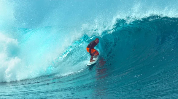 CLOSE UP: Focused pro male surfer riding a powerful big barrel wave in beautiful tropical Tahiti. Spectacular shot of young extreme sportsman riding a deep blue tube ocean wave on his surfboard.