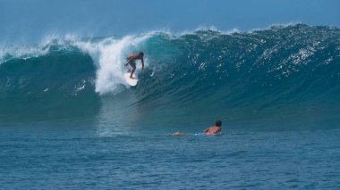 Yakın: Genç erkek sörfçü arkadaşı öğleden sonra güneşin altında ışıltılı bir büyük zümrüt namlu okyanus dalga binmek izliyor. Pro surfboarders epik tüp dalgaları sürme tarafından'nda kendi yaz tatil keyfini