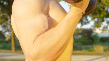 Yakın: bir demir dumbbell sakin yeşil park curling tarafından güçlü kollarıyla egzersiz yaparak bilinmeyen genç kaslı adam. Doğada bukleler yapıyor uygun tanınmaz erkek vücut yaz güneş parlar.