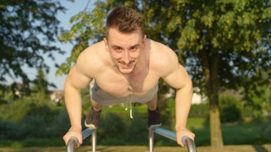 Portre: paralel bar serin açık spor salonunda egzersiz genç adam gülümsüyor. Gönderme yapan neşeli kaslı adam ups güneşli bir günde bir büyük egzersiz parkta. Açık havada atletik erkek eğitim.