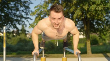 Portre: itme yapıyor atletik genç beyaz adam paralel bar yeşil park ups. Gülümseyen yetişkin erkek atlet eğleniyor yaz aylarında açık spor salonu içinde çalışma. Dışarıda egzersiz kas adam.