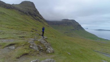 Anteni: genç kadın dik bir çimenli tepeden muhteşem sahilden bulutlu bir günde Faroe Adaları güzel kırsal kesimde fotoğraf çekme el değmemiş dağlarla çevrili. Kadın fotoğrafçı
