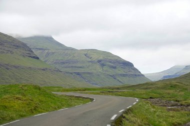 Bulutlu gökyüzüne doğru yükselen görkemli yüksek dağlar arasında çalışan boş asfalt yol manzarası. Nefes kesen manzaralı yoldan Faroe Adaları'çarpıcı engebeli yatay geçiş.