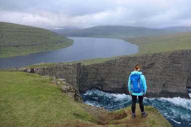Hava: Genç kadın gezgin için nefes kesici yeşil peyzaj moody Faroe Adaları'nda görmek için bir uçurumun kenarında zamları. İskandinavya hiking kadın hala göl ve dalgalar kayalara çökmesini gözlemler
