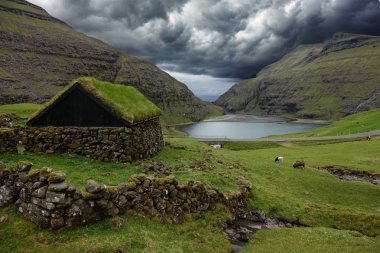 Uçsuz bucaksız vadide çimlerle kaplı geleneksel İskandinav evleri. Faroe Adaları 'ndaki küçük bir köyün her iki tarafında yükselen dik dağların nefes kesici görüntüsü.