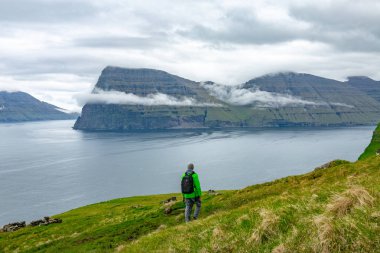 Bilinmeyen bir adam kayalıklarla ve sakin okyanus bulutlu bir günde Faroe Adaları bakan sarp bir çimenli tepe boyunca zamları. Engebeli İskandinav kıyı ve çok yüksek yeşil dağların Breahtaking görünümünü.