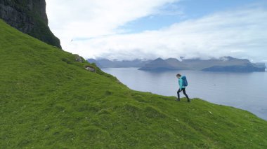 Hava: Kadın turist okyanusun mükemmel manzaralı çimenli tepeye üzerinde soğuk bir sabah zamları. Engin deniz ve yeşil anakara bakan dik bir dağa yolculuk hiking üzerinde genç kadın yürüyor.