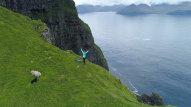 AERIAL, COPY SPACE: Cheerful woman standing on the edge of a cliff with outstretched arms while looking at the ocean. Female tourist is happy after hiking up a steep grassy mountain in Faroe Islands. clipart