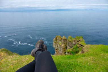 POV: bir uçurum ve aşağıda sakin okyanus bakarak çimenli kenarında oturuyor. Faroe Adaları dağlarda rahatlatıcı kadında tozluk ve yürüyüş botları. Pitoresk İskandinav shore.