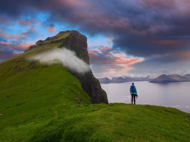 Kadın fotoğrafçı bir uçurumun kenarında duruyor ve muhteşem mavi ve turuncu akşam gökyüzünde izler. Çarpıcı görünümü muhteşem yeşil kayalıklarla ve pitoresk Faroe Adaları dağlarda.