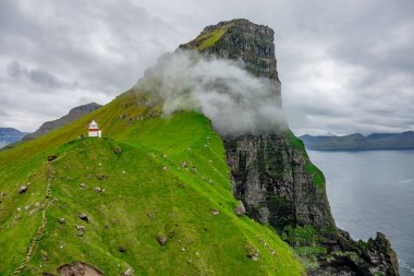 AERIAL: Flying towards a small white lighthouse on top of the grassy hill in Faroe Islands. White clouds drift along the rocky cliffs on a picturesque fjord in the scenic Scandinavian wilderness. clipart