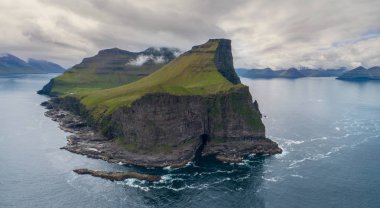 Hava: kayalık bir siyah uçurum yakın çimenli bir tepenin üstüne oturan küçük beyaz deniz feneri bulutlu gün sakin deniz görünüyor. Bir küçük yeşil adacık Faroe Adaları'nın nefes kesen havadan görünümü.