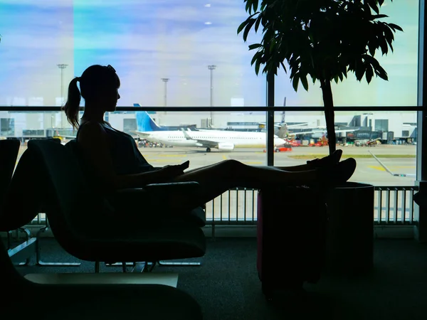 Siluet: bankta oturan ve onun tatil beldesi onun uçuş için havaalanında bekleyen kadın. Tanınmayan kadın turist Uluslararası Havaalanı terminal büyük pencereden bakıyor.