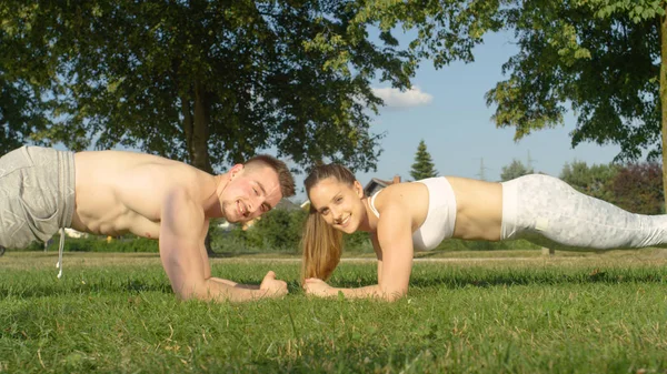 PORTRAIT: Cheerful young training partners work out their abs in the tranquil park on a sunny summer day. Happy fit Caucasian couple exercising together in the outdoors and holding a plank position.