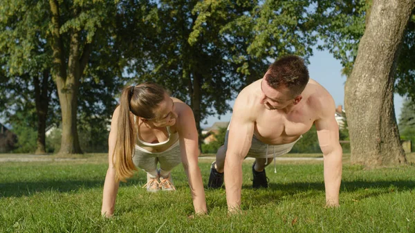 Yakın: Genç gülümseyen adam yerleştirmek ve kadın basın ups ve açık havada yaz güneşte egzersiz. Atletik kız ve kaslı erkek arkadaşı güneşli parkta birlikte çalışma keyfini çıkarın. Çift eğitim