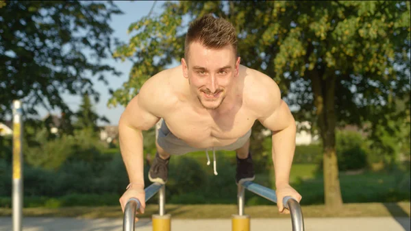 Portre: itme yapıyor atletik genç beyaz adam paralel bar yeşil park ups. Gülümseyen yetişkin erkek atlet eğleniyor yaz aylarında açık spor salonu içinde çalışma. Dışarıda egzersiz kas adam.