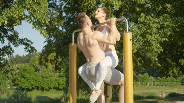 Yakın: Güçlü beyaz çift çekme işi uygun ups harika bir açık spor salonunda birlikte. Mutlu onun kaslı erkek mu çene ups metal çubuğu yeşil parkta kız bacakları ile sarılı.